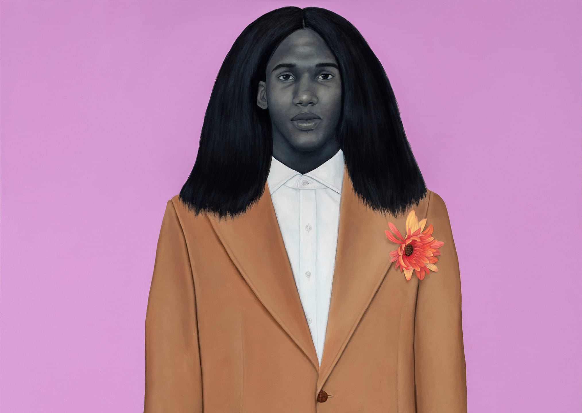 A person with long black hair in a tan suit with a flower pinned, standing against a pink background.
