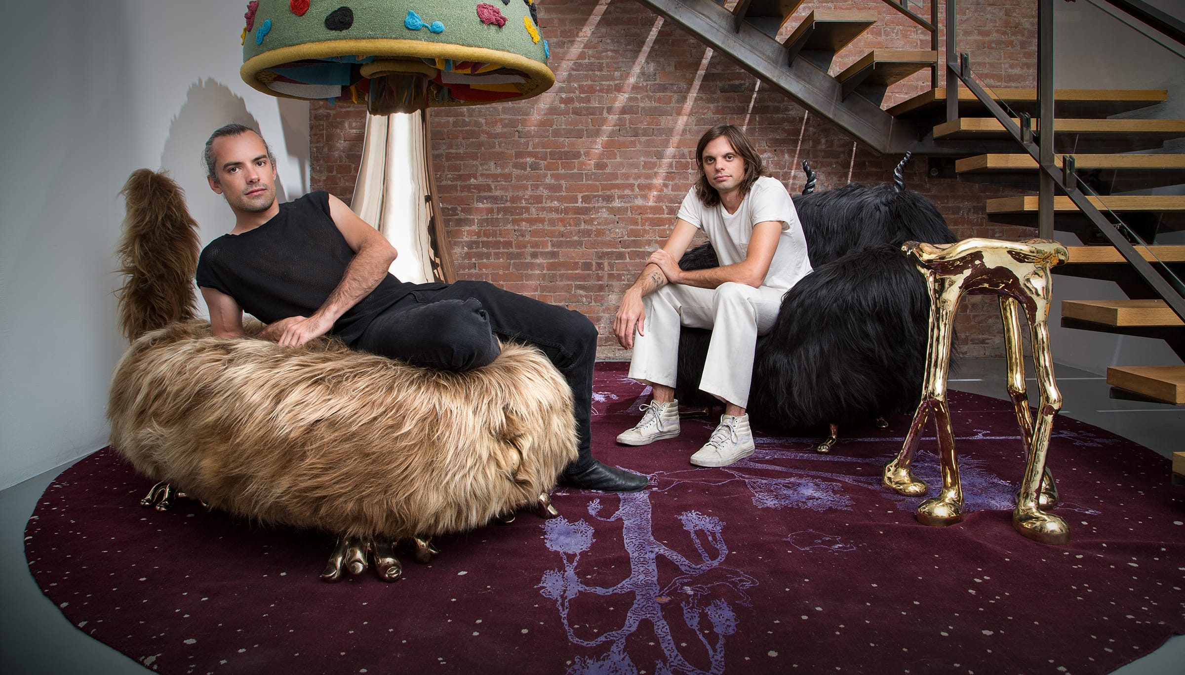 Two people sitting on furry chairs under a large mushroom lamp in a modern room with a brick wall and a staircase.