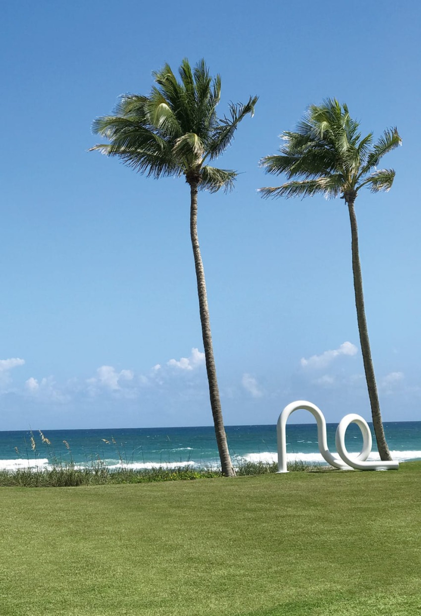 Two palm trees sway by the ocean with a white sculpture, under a clear blue sky.