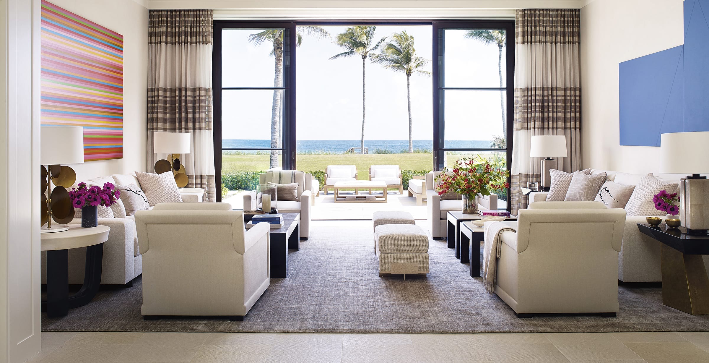 Spacious modern living room with white sofas, vibrant artwork, ocean view through large windows, and palm trees outside.