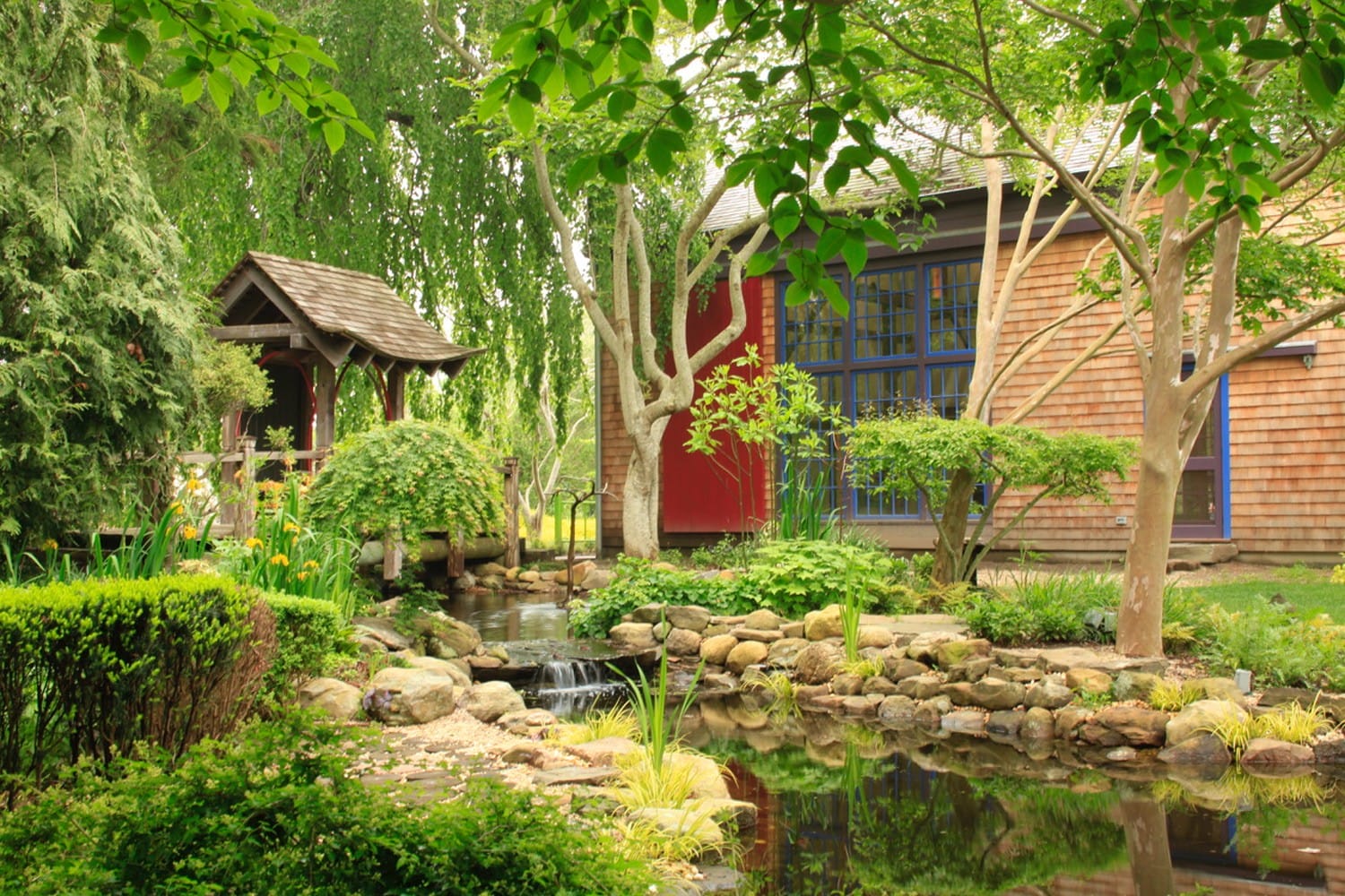 Garden landscape with a small pond, wooden footbridge, and rustic house surrounded by lush greenery and trees.