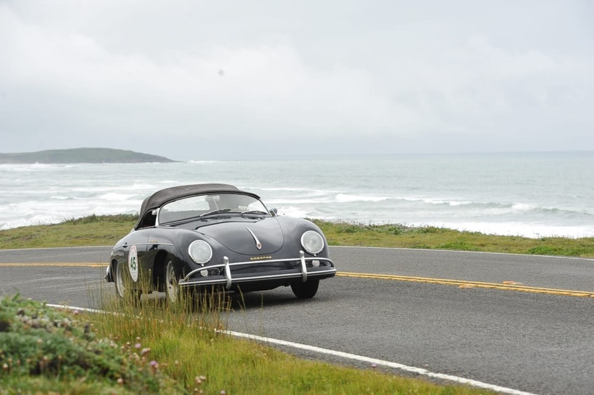Vintage black convertible driving on a scenic coastal road with ocean waves in the background on a cloudy day