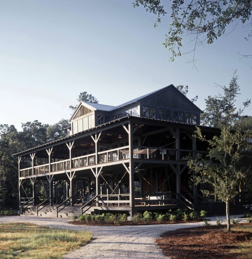 Rustic two-story wooden cabin with a wraparound porch surrounded by trees and a gravel path in the foreground.
