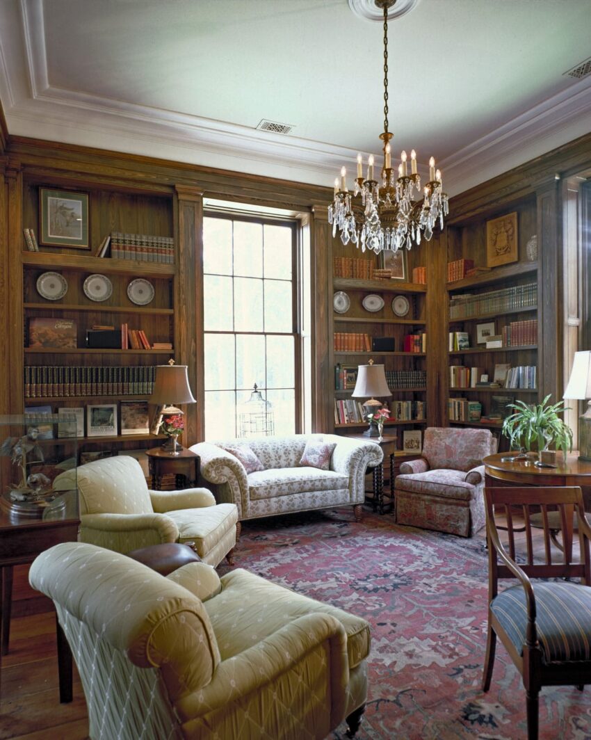 Cozy library room with armchairs, sofa, chandelier, wooden bookshelves filled with books, and a large window.