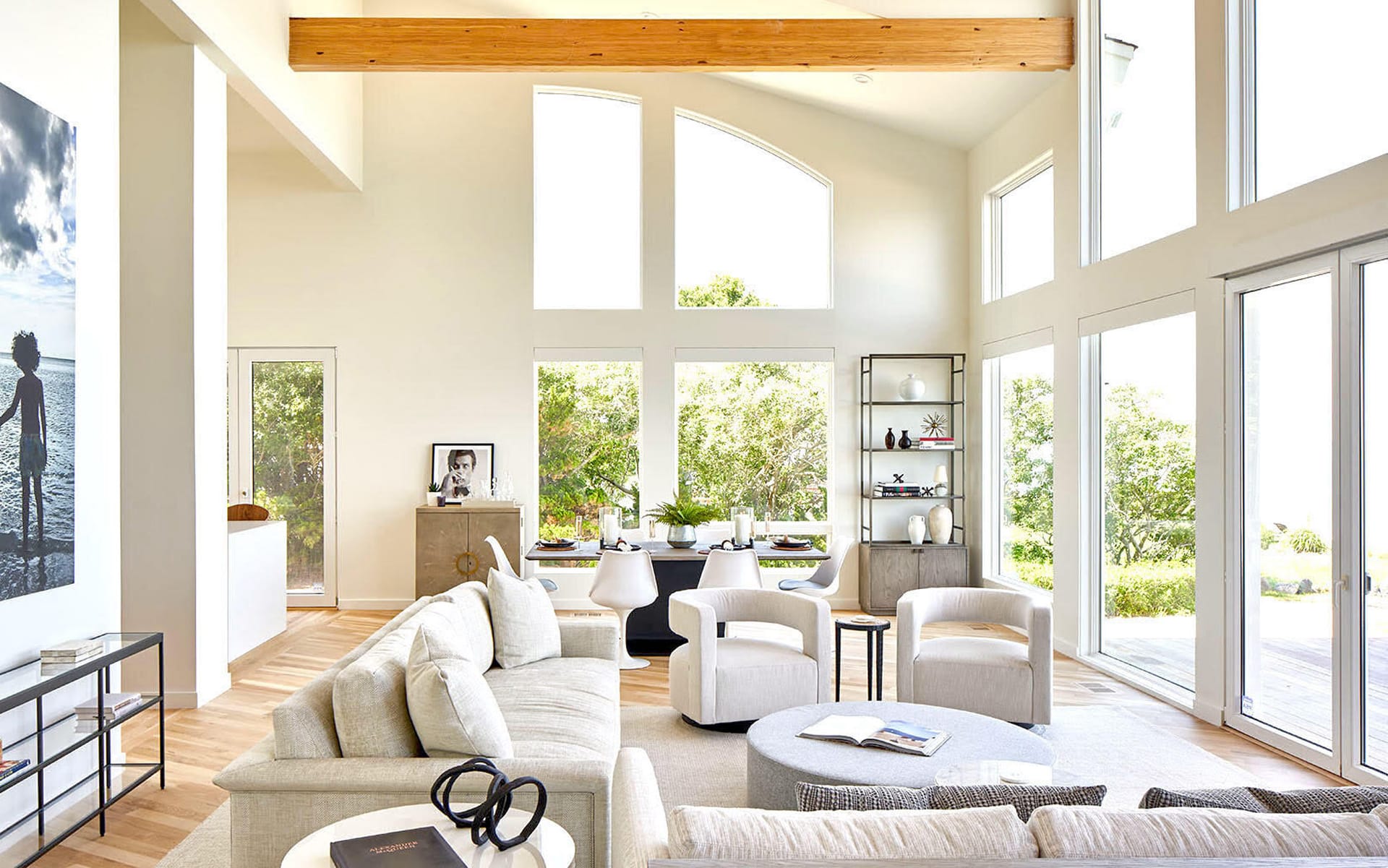 Modern living room with large windows, light furniture, and wooden beam accentuating natural light and greenery outside.