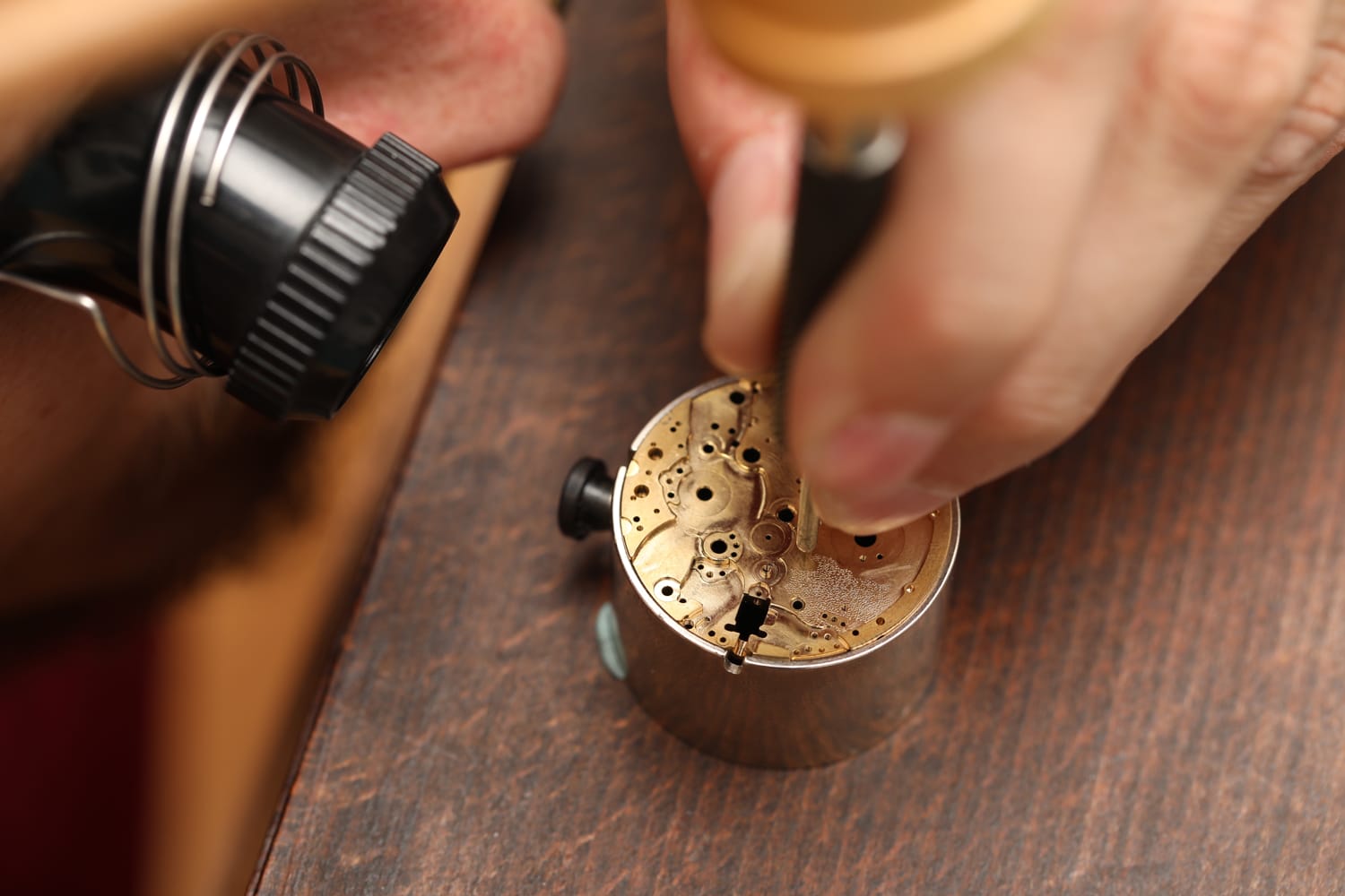 Close-up of person repairing a watch mechanism with a small tool on a wooden table.