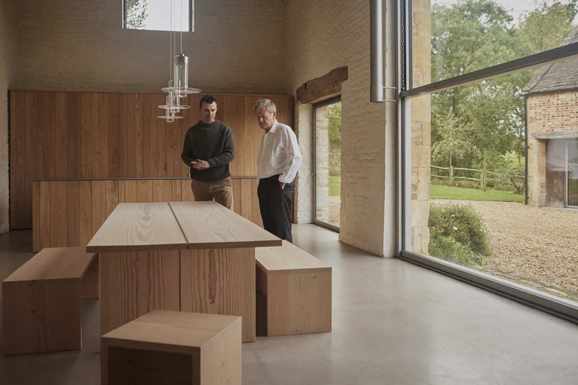 Two people standing in a minimalist dining room with wooden furniture and large window views of a garden outside.