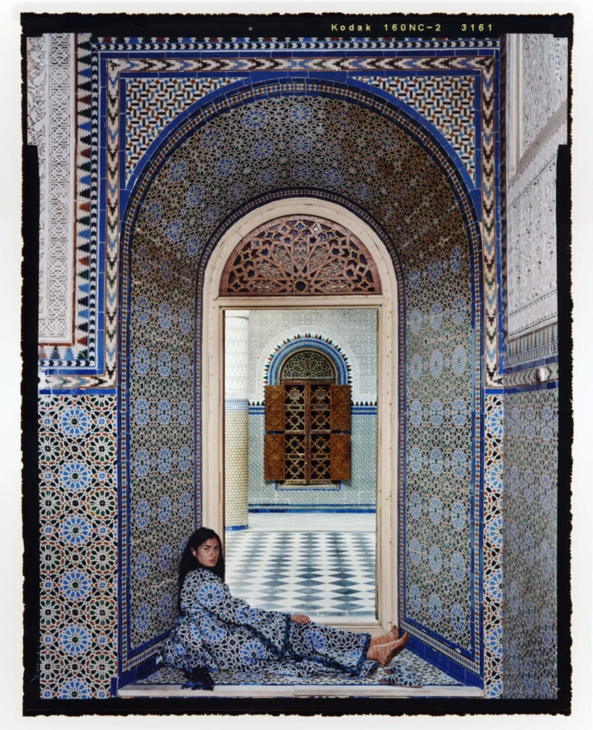 A person in patterned attire sits by an ornate archway in a tiled room with intricate blue and white designs.