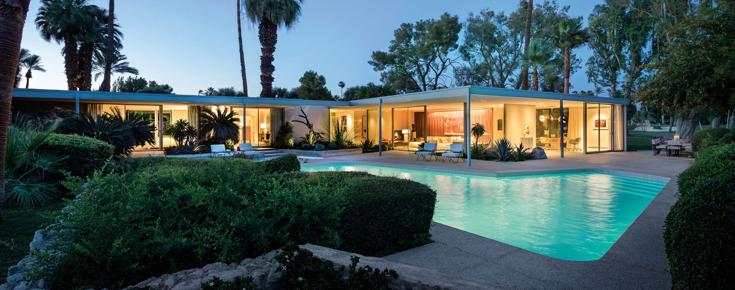 Modern glass house with a glowing pool, surrounded by palm trees and greenery, under a twilight sky.