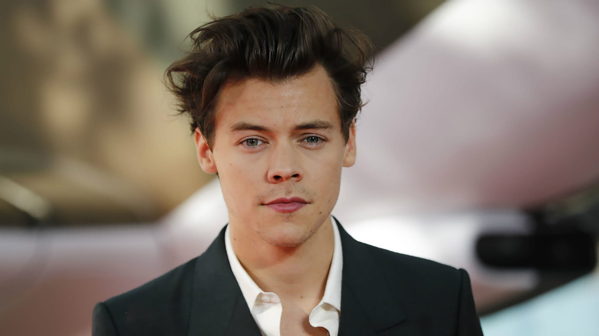 A person with short dark hair wearing a black jacket and white shirt, standing in front of a blurred background.