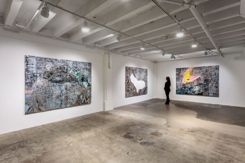 Person admiring abstract art in a modern gallery with three large paintings displayed on white walls and concrete floor.