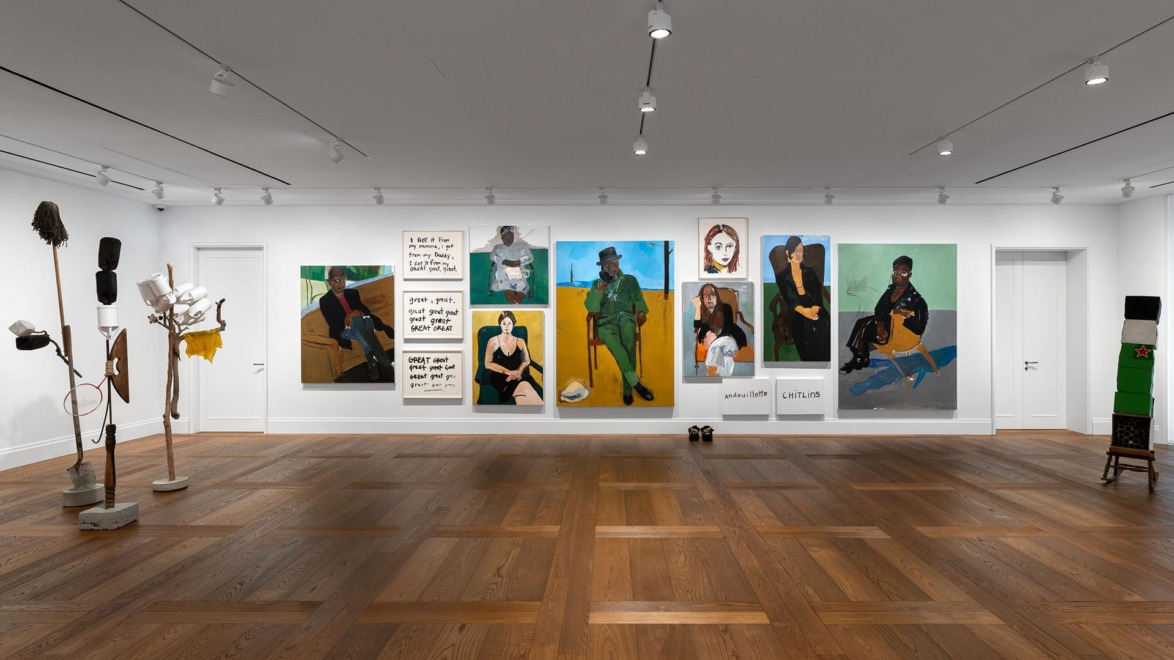 Art gallery with various colorful portraits on the wall and sculptural installations on wooden floor in a well-lit room.
