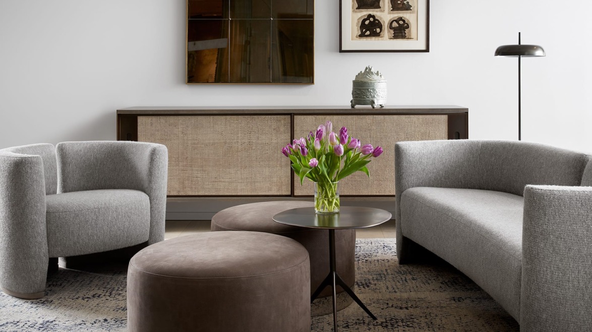Modern living room with gray curved sofa and armchair, round brown ottoman, and a vase of purple tulips on a small table.