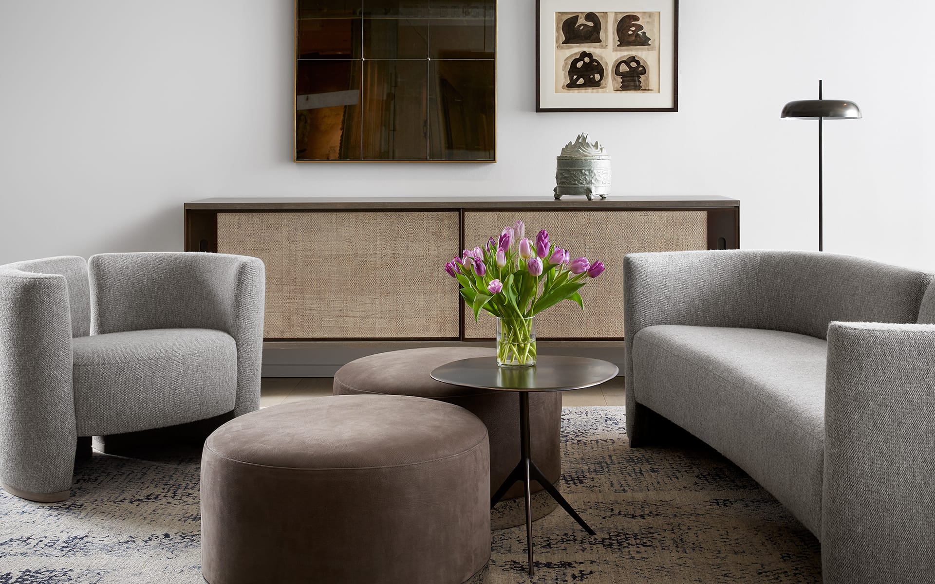 Modern living room with gray curved sofa and armchair, round brown ottoman, and a vase of purple tulips on a small table.