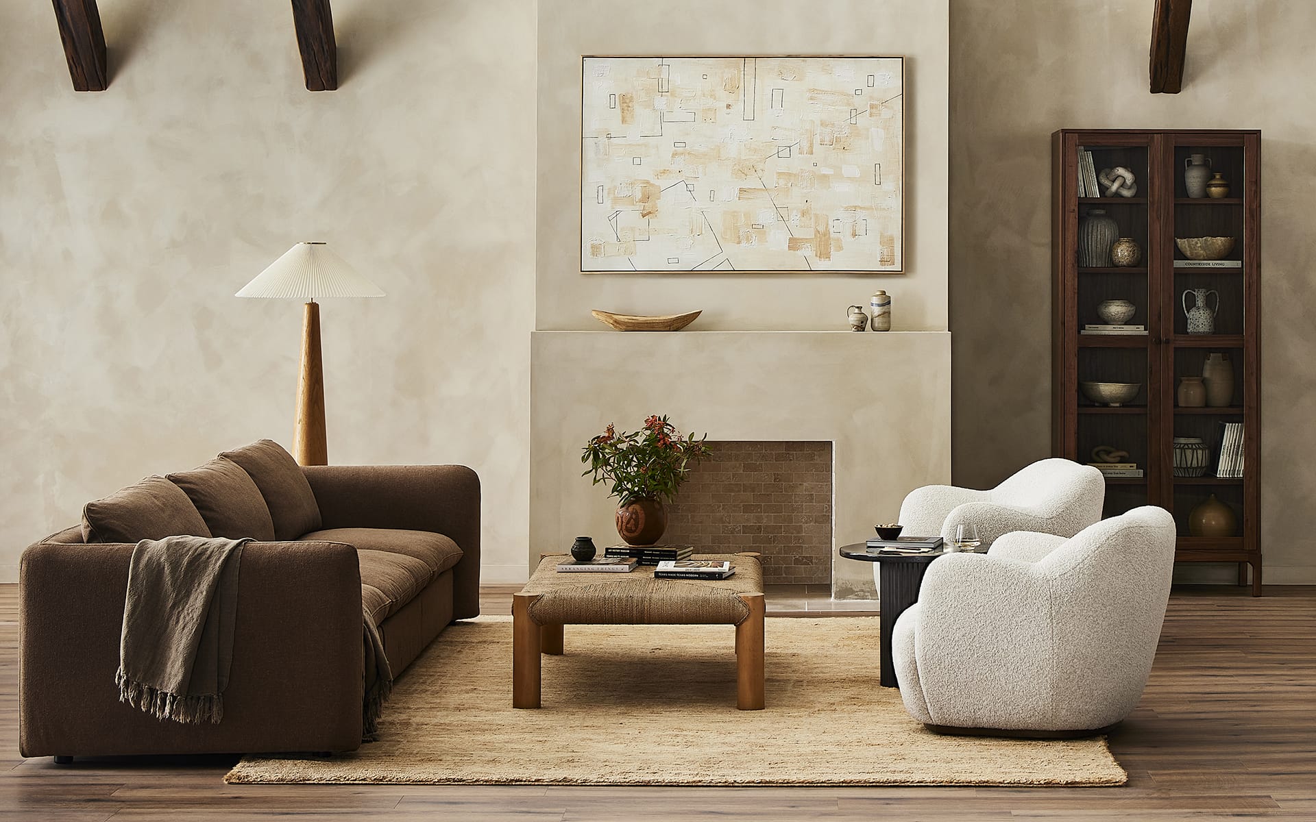 Modern living room with brown sofa, two white armchairs, coffee table, lamp, abstract art, and bookshelf on wooden floor.
