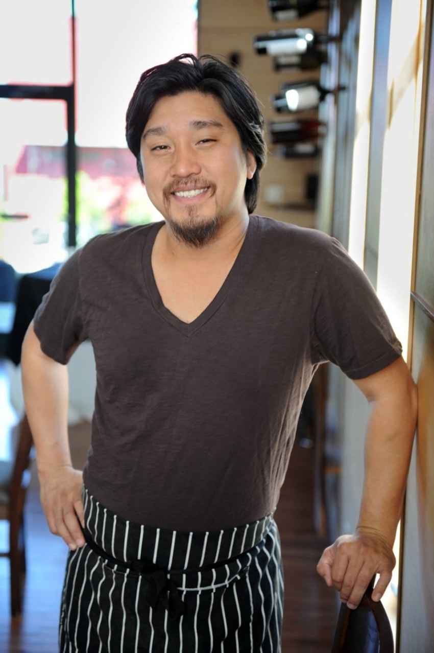 Smiling chef wearing a black apron stands with hands on hips in a bright restaurant interior.