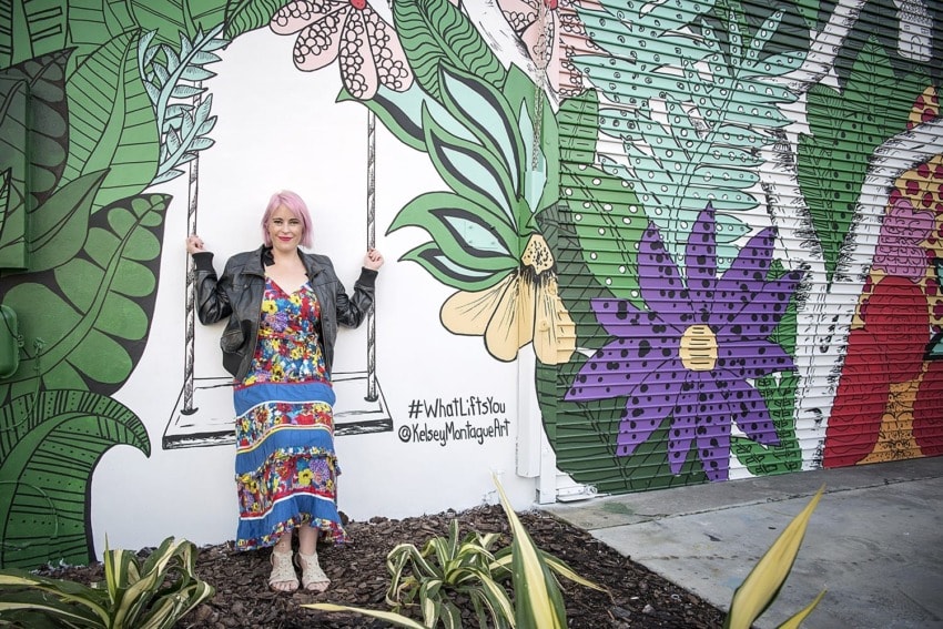 Person with pink hair in a colorful dress sitting on a swing in front of a vibrant mural with flowers and leaves.