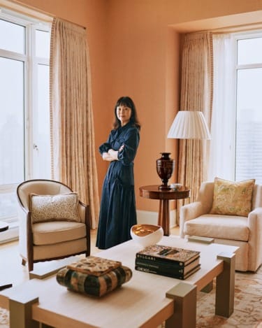 A person standing in a stylish living room with modern furniture, large windows, and a warm color scheme.