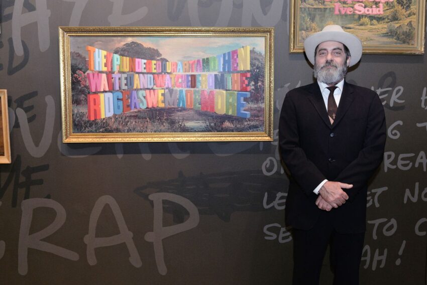 Man in a suit and hat standing next to a framed colorful text art piece on a wall.