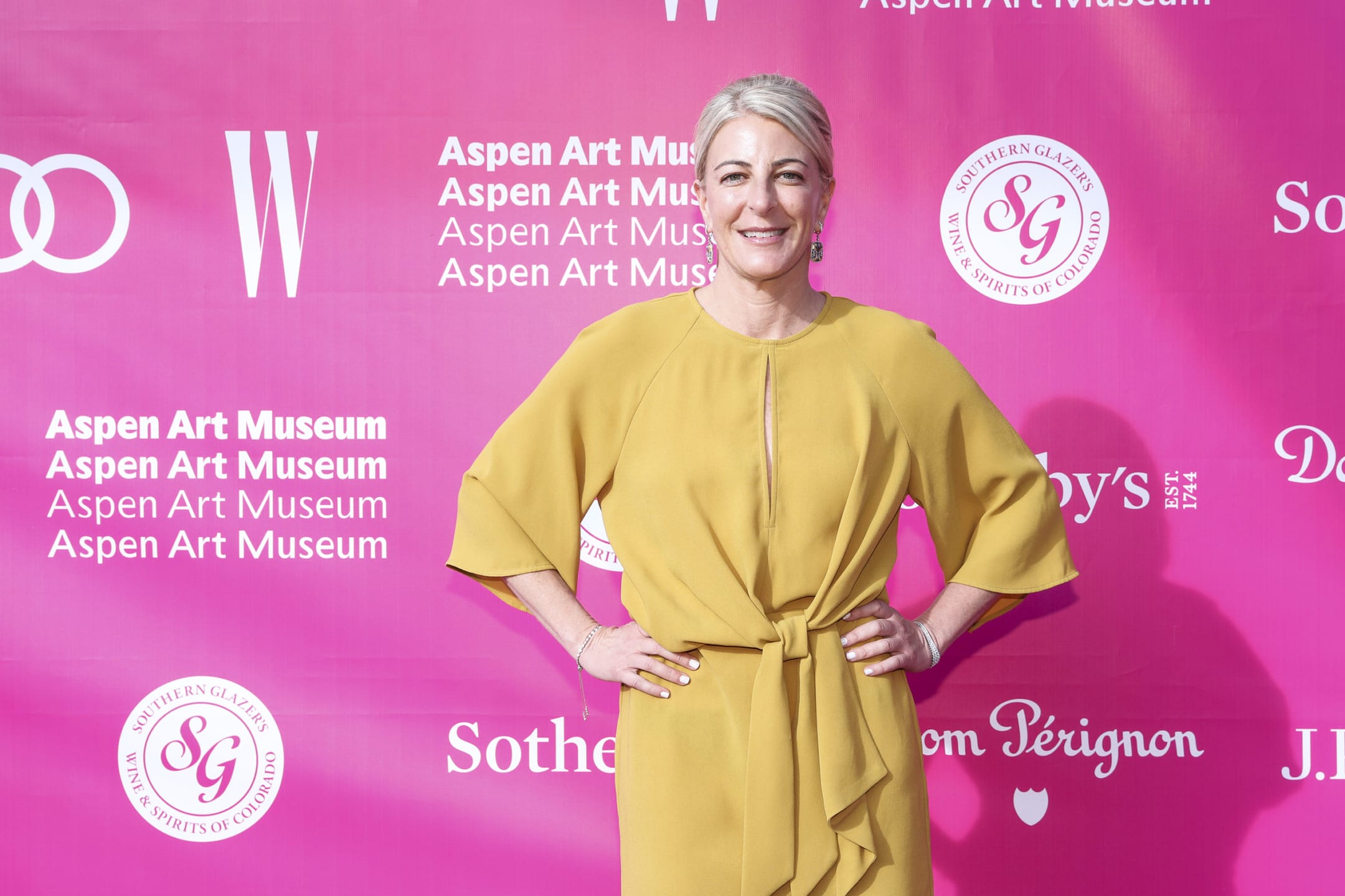 Person in a yellow dress posing on a pink event backdrop with various sponsor logos and text.