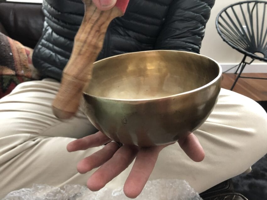 Person holding and playing a brass singing bowl with a wooden mallet in a cozy indoor setting.