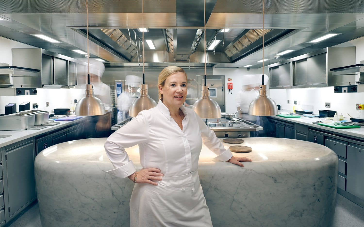 Chef standing confidently in a modern, stainless steel kitchen with marble counter and pendant lights.