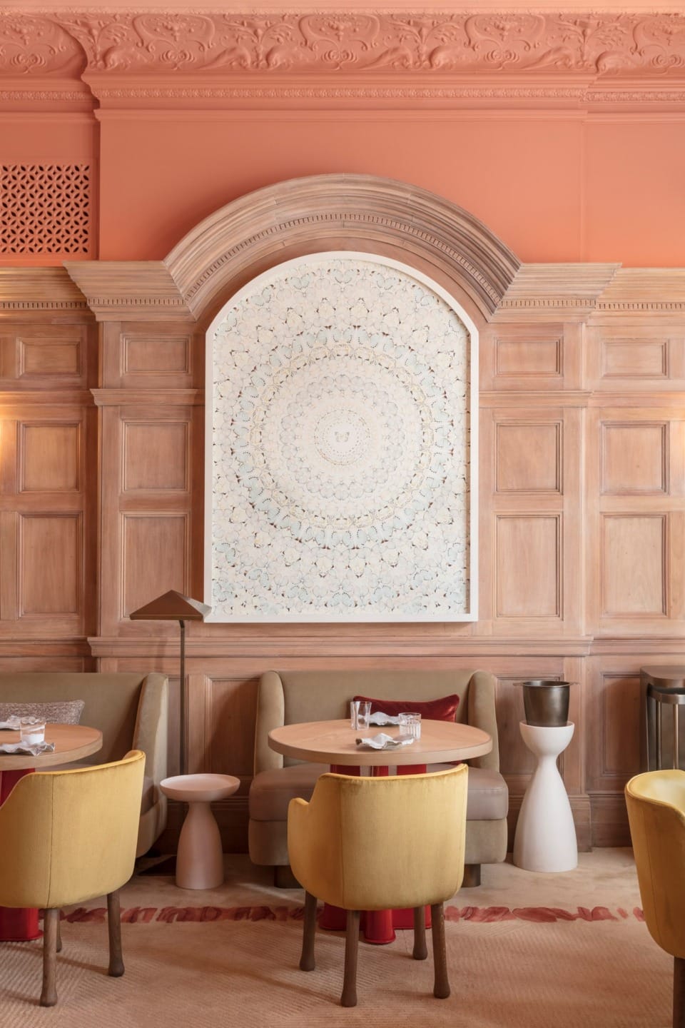 Elegant restaurant interior with light wood paneling, peach walls, yellow chairs, and a large circular patterned artwork.