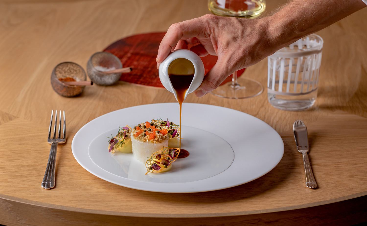 Hand pouring sauce over elegantly plated gourmet dish on a white plate with cutlery and a glass of water on a wooden table.