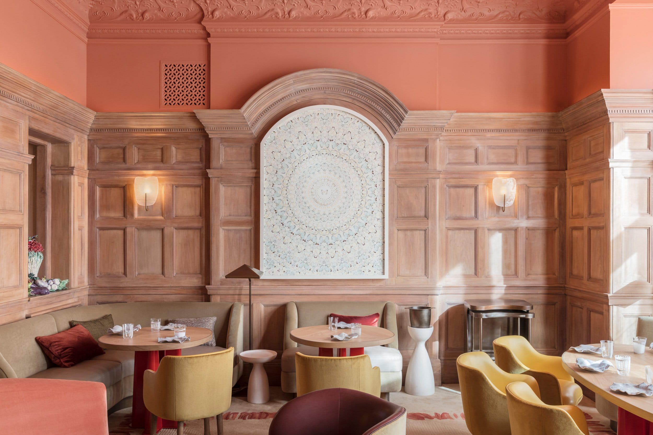 Elegant restaurant interior with ornate wood paneling, round tables, colorful chairs, and a decorative wall art centerpiece.