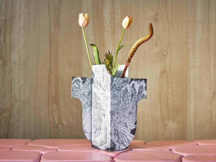 Modern vase with sculpturesque design holding tulips and a leafy plant on a pink surface against a wooden background.