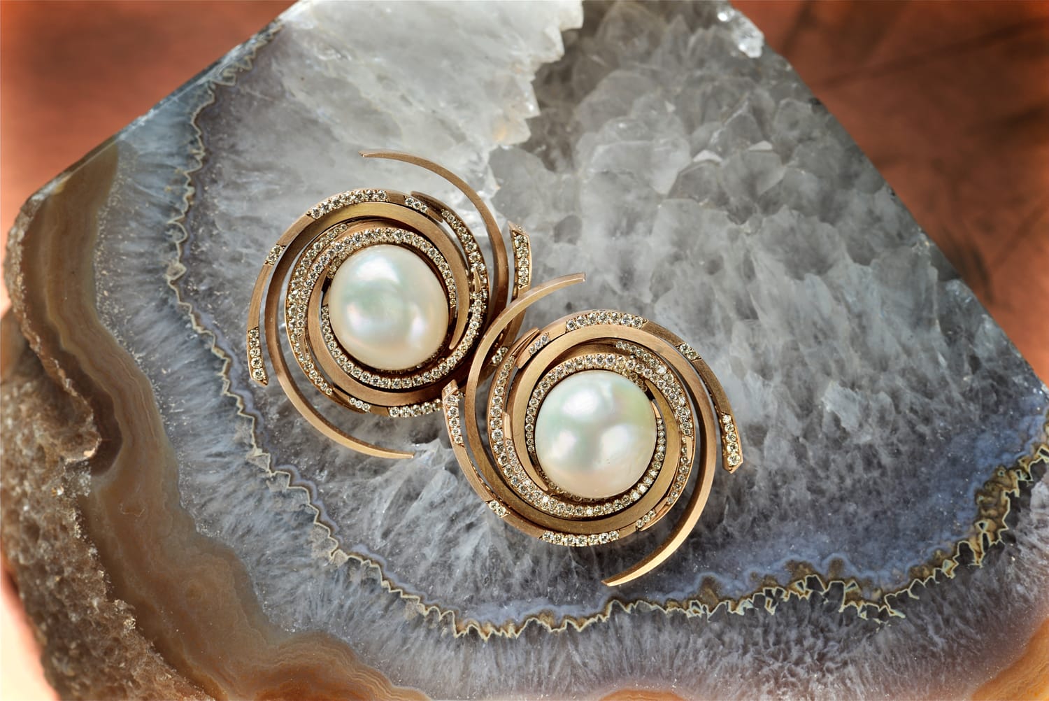 Gold swirl earrings with pearls and diamonds displayed on a textured stone surface.