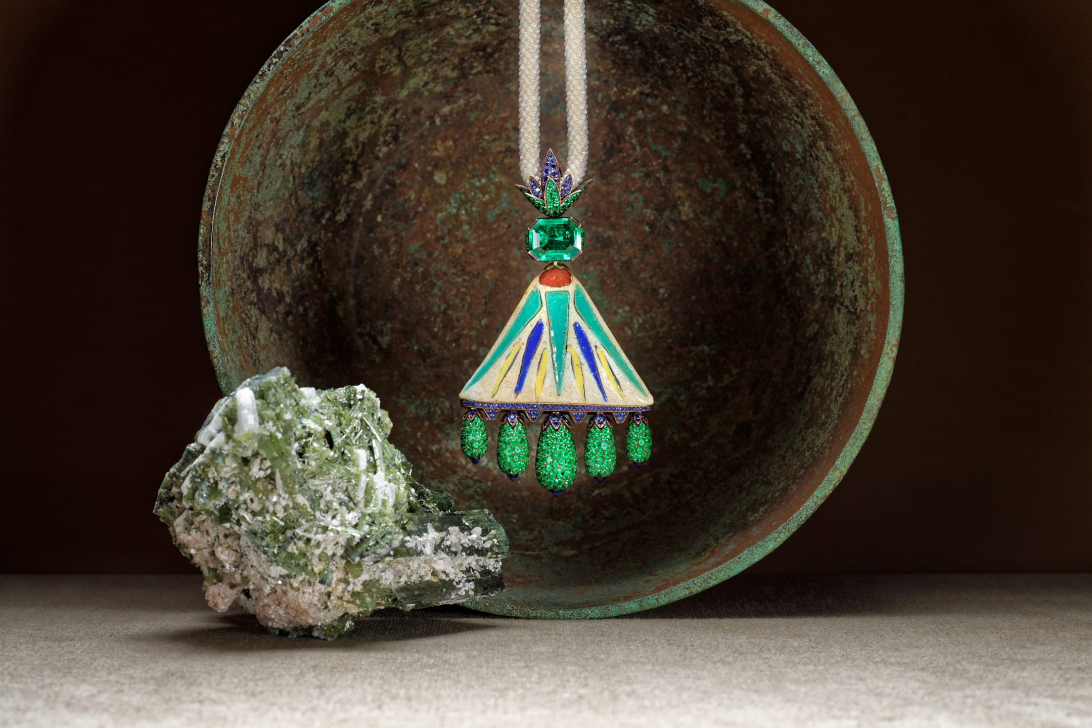 Colorful, ornate pendant with green gemstone detail hanging in front of a textured, rusted bowl and a green mineral rock.
