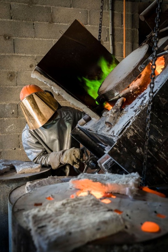 Worker in protective gear pours molten metal into a mold in an industrial foundry with a green flame visible.