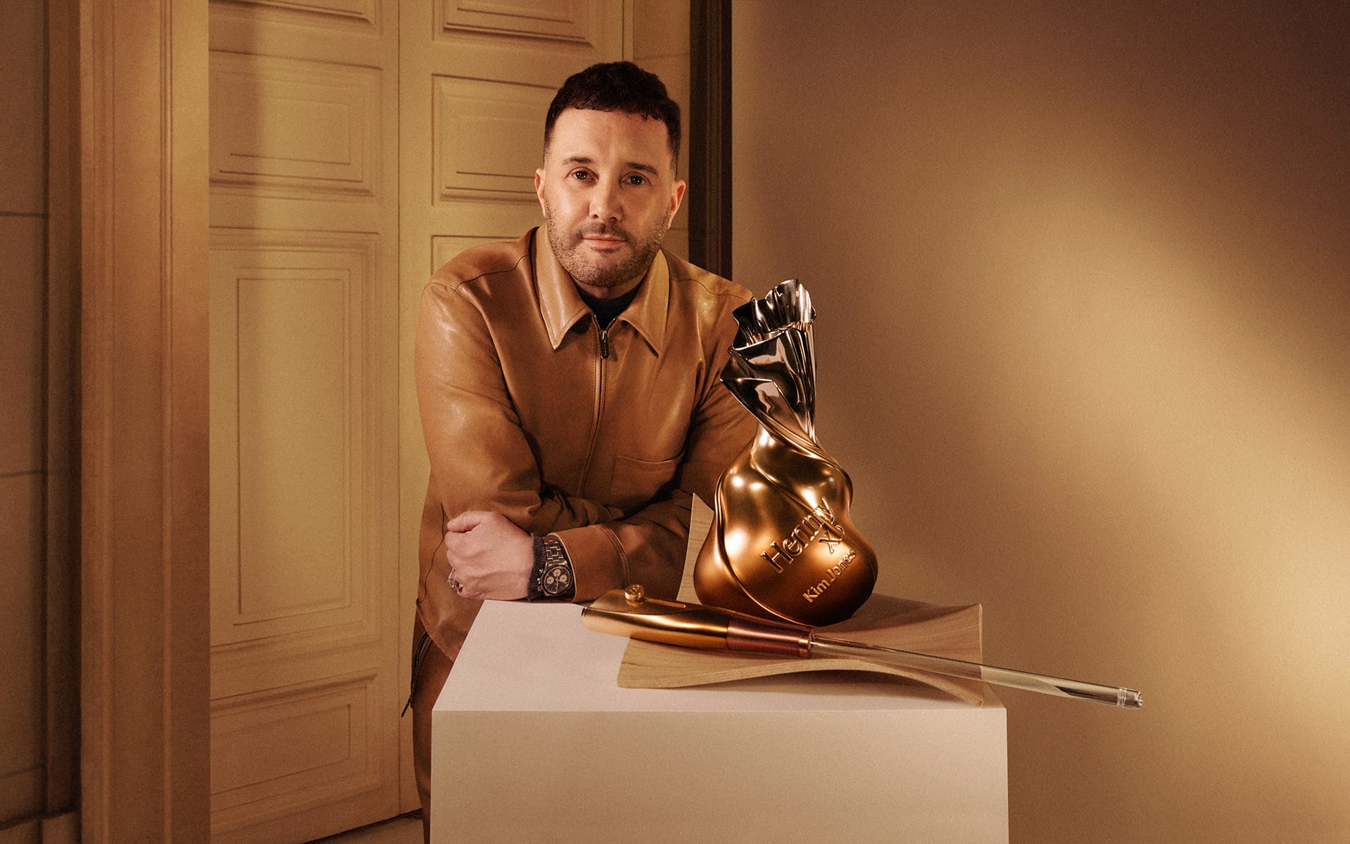Man in a leather jacket with a golden trophy and metal sword on a pedestal in an elegantly lit room.