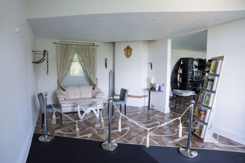 Modern living room with patterned floor, white sofa, glass table, and bookshelves, roped off for display.
