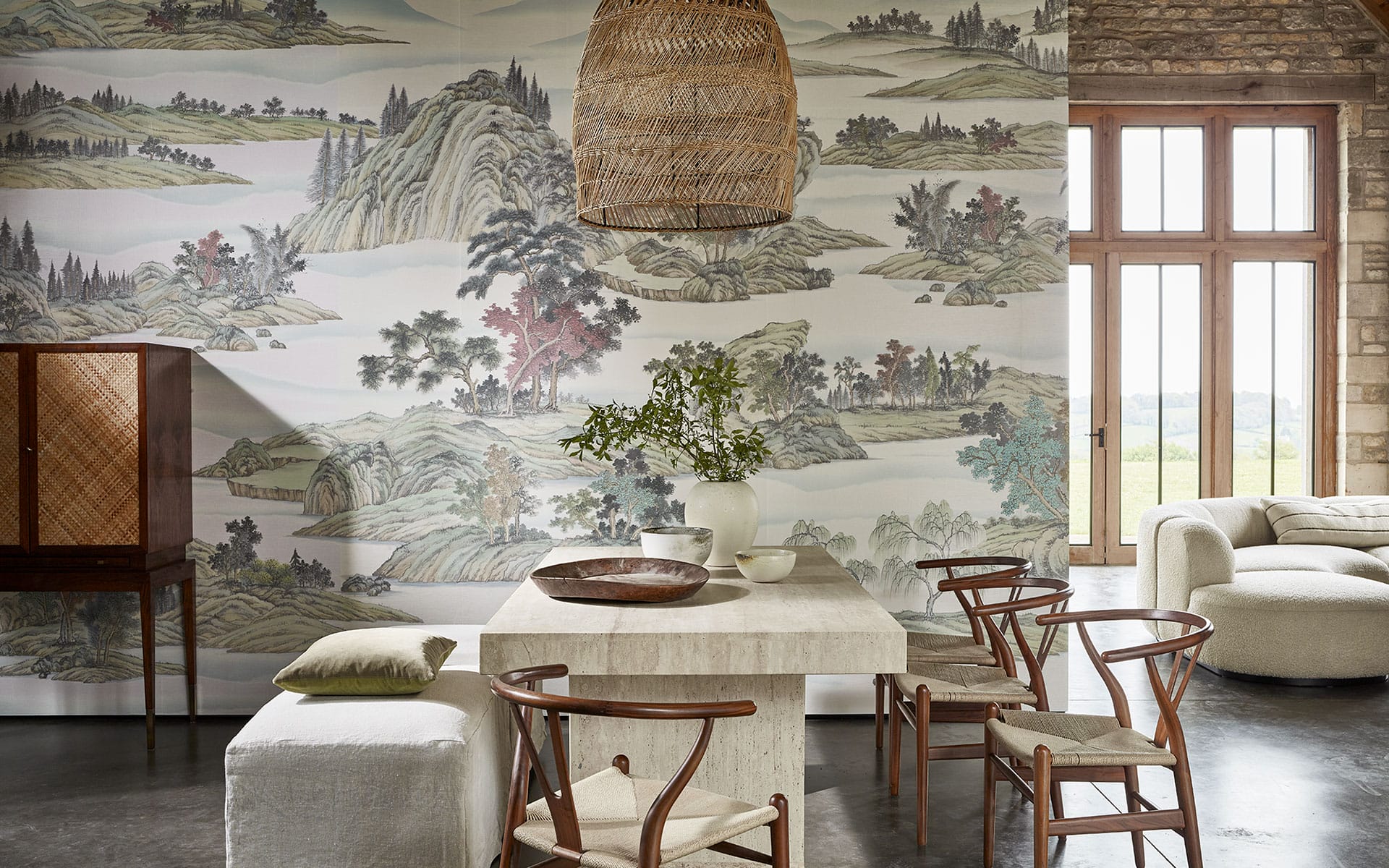 Dining area with a wooden table and chairs, decorative wallpaper, a woven pendant light, and large windows in a cozy room.