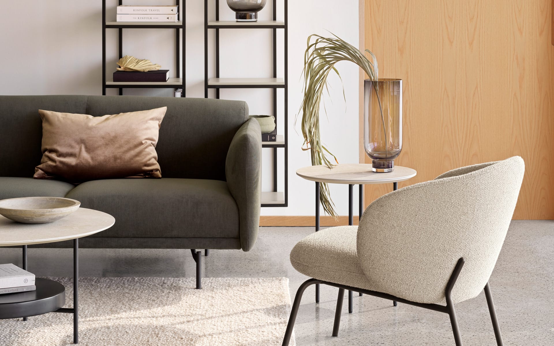 Modern living room with a green sofa, beige armchair, round tables, decorative vase, and bookshelves.