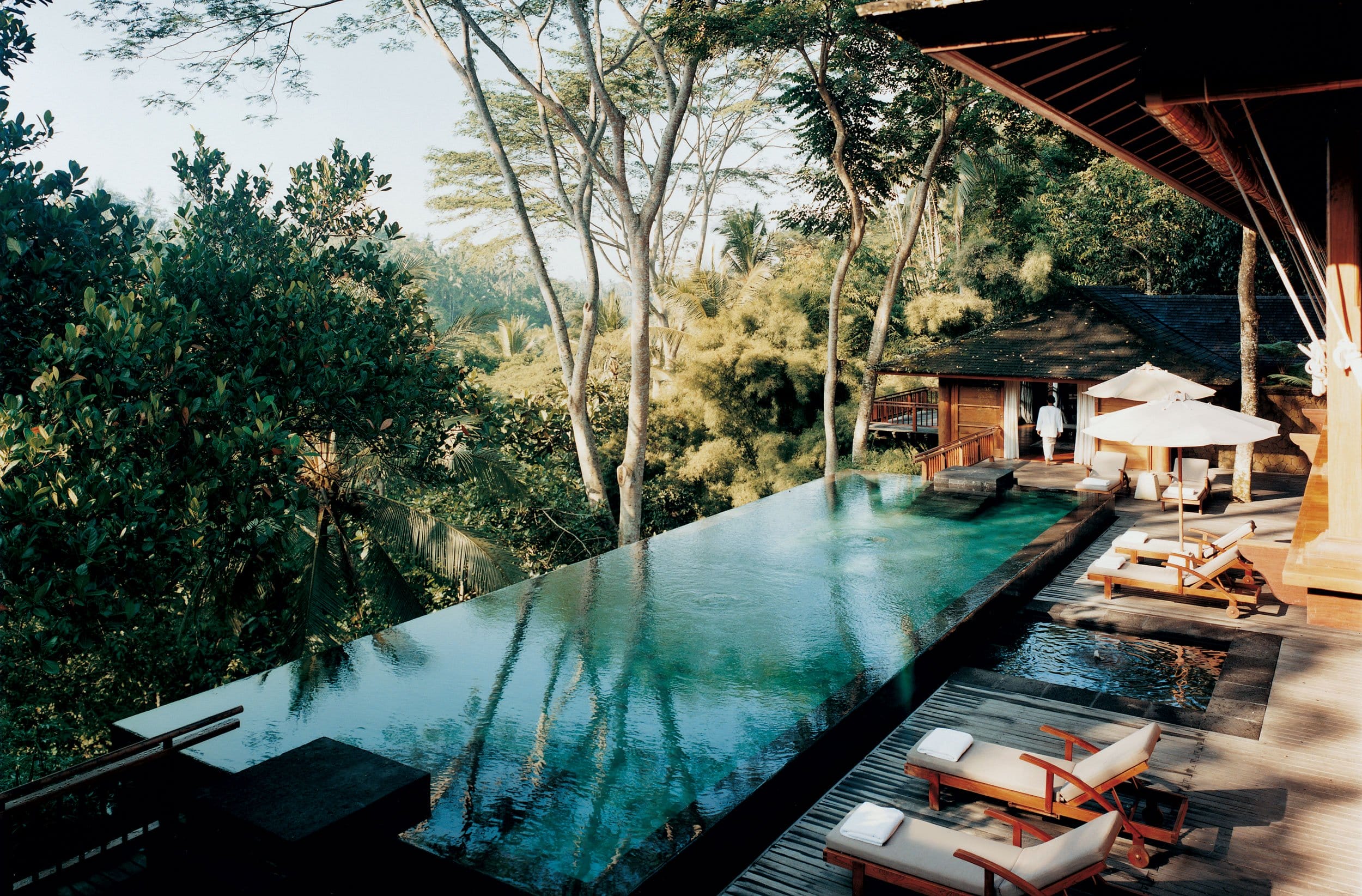 Luxurious outdoor infinity pool overlooking lush greenery surrounded by lounge chairs and umbrellas at a tropical resort.