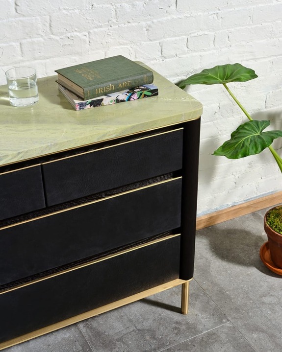 Black dresser with green marble top, books, a glass of water, and a potted plant beside a white brick wall.