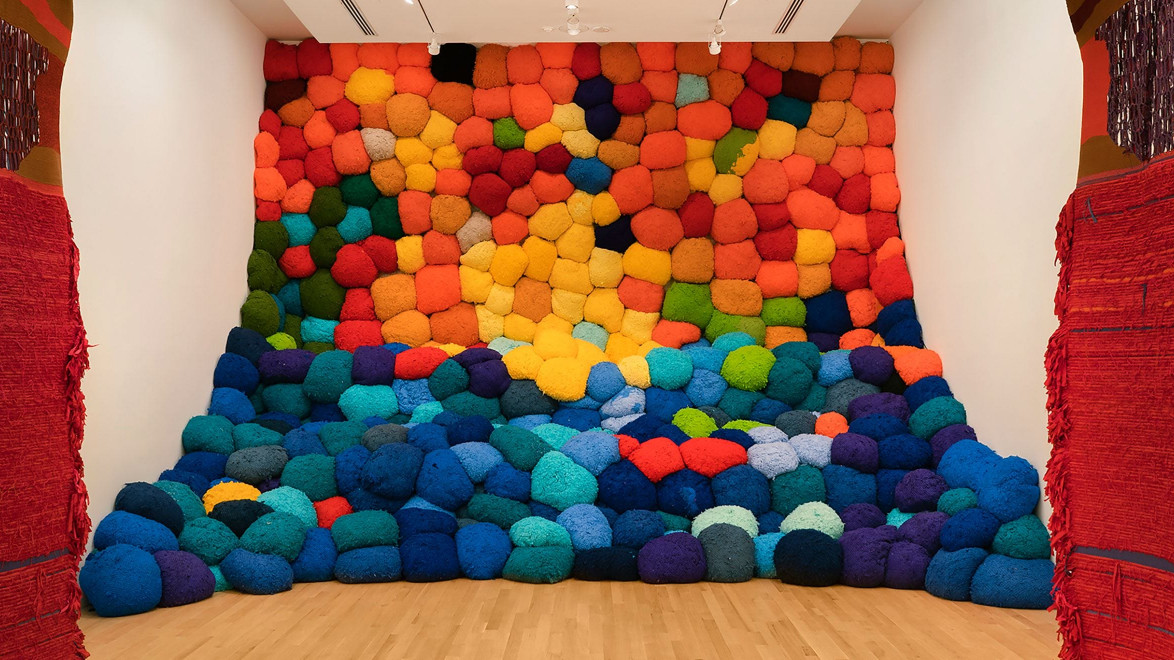 Colorful installation of large, fluffy cushions stacked against a wall, creating a gradient from warm to cool tones.