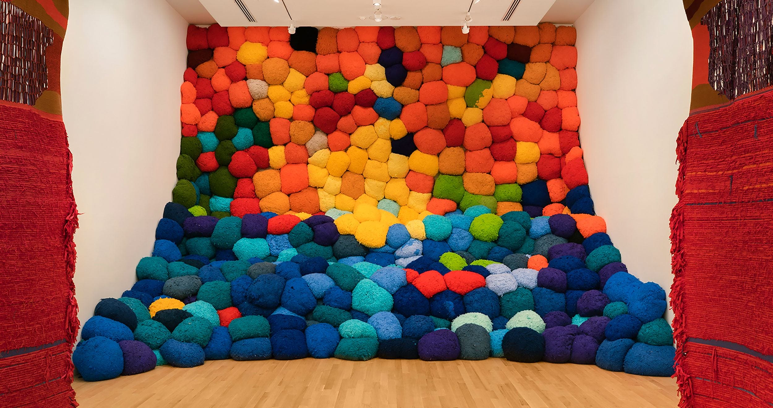 Colorful installation of large, fluffy cushions stacked against a wall, creating a gradient from warm to cool tones.
