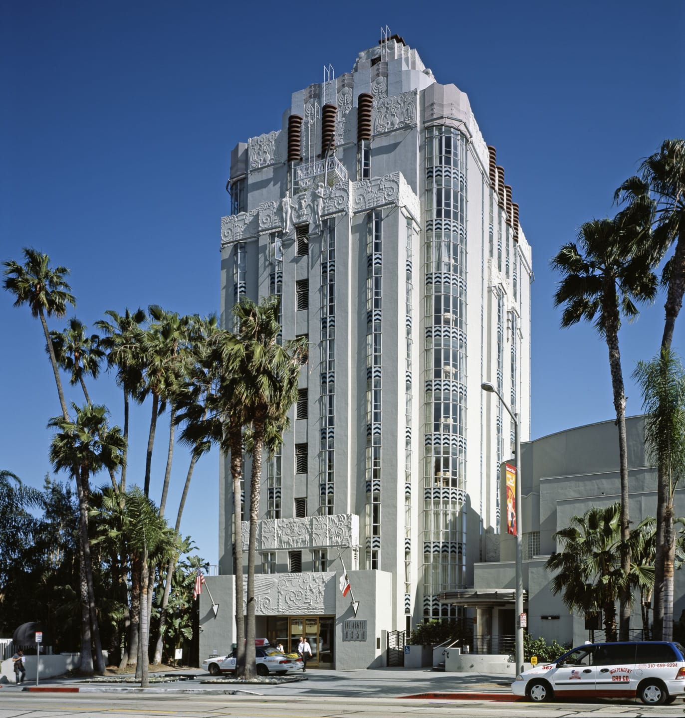 Art Deco building with detailed facade, palm trees in foreground, clear blue sky, and cars parked along the street.
