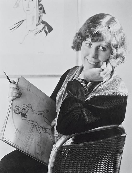 Smiling woman seated, holding a sketchbook with an illustration, framed art visible in the background.