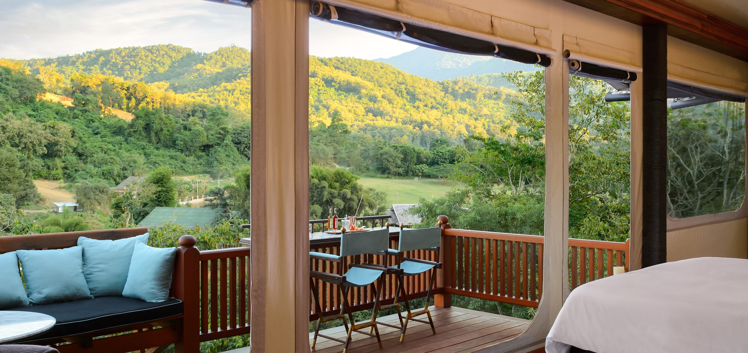 Mountain view from a cozy room's balcony with seating area, surrounded by lush greenery and afternoon sunlight.