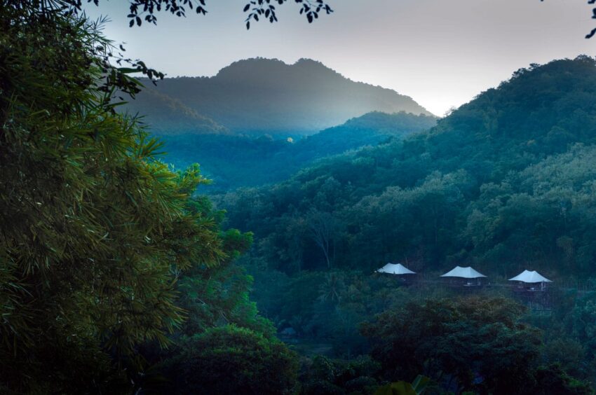 Tents nestled among lush green hills with a mountain in the background during sunrise.