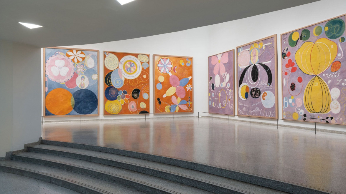 Colorful abstract paintings displayed on a white gallery wall with a curved staircase in the foreground.