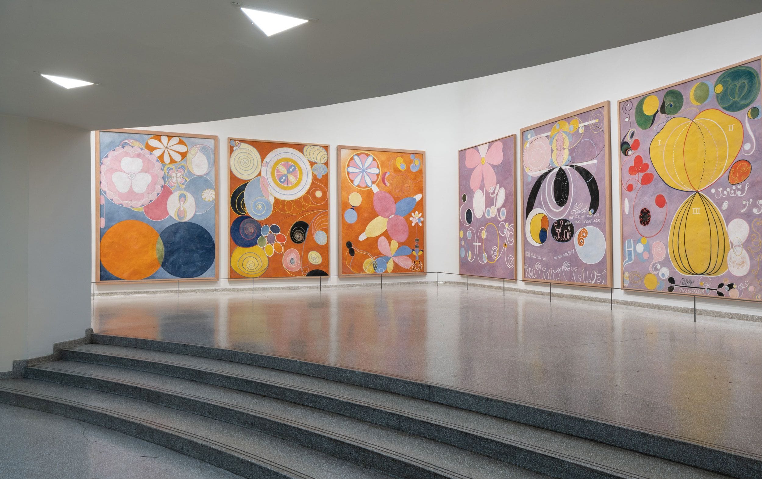 Colorful abstract paintings displayed on a white gallery wall with a curved staircase in the foreground.
