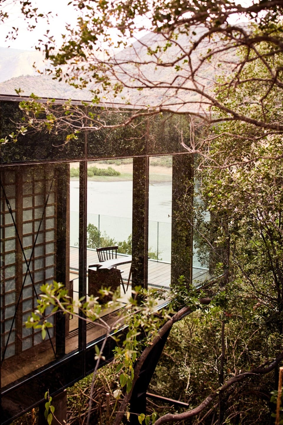 Modern glass house with balcony surrounded by trees overlooking a serene river scene.