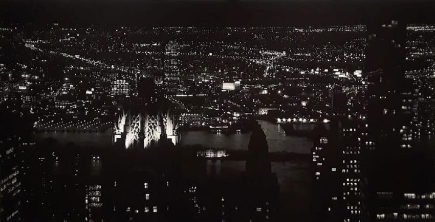 Aerial view of a city skyline at night with illuminated buildings and a river in the background.