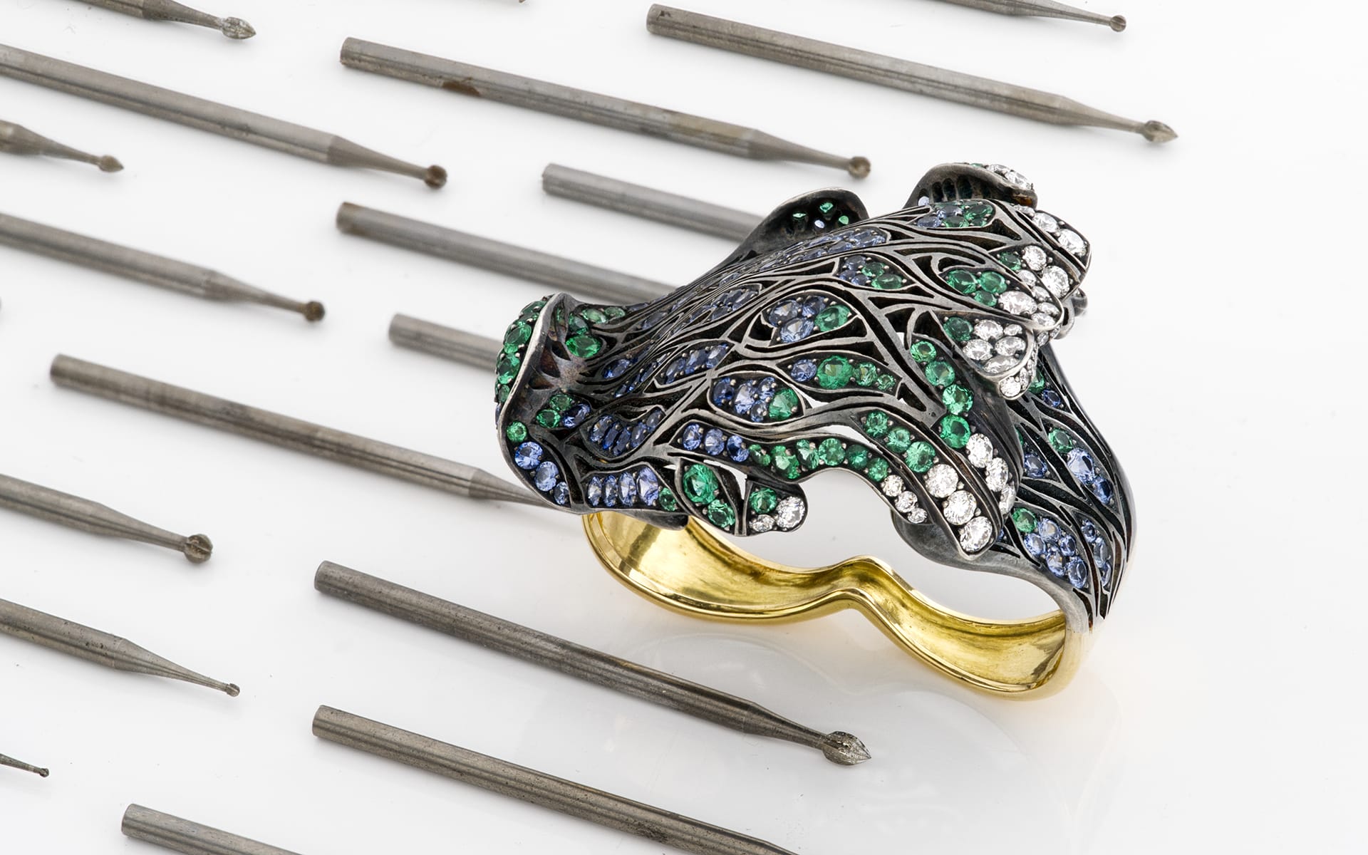 Intricate silver and gold ring with green, blue, and clear gemstones, surrounded by metal engraving tools on a white surface.
